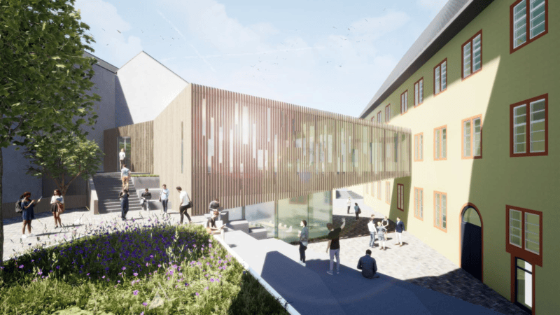Moderner, langgestreckter Baukörper mit vertikalen Lamellen zwischen zwei historischen Gebäuden, umgeben von gepflastertem Hof und Grünfläche mit Blumen, mehrere Personen im Außenbereich