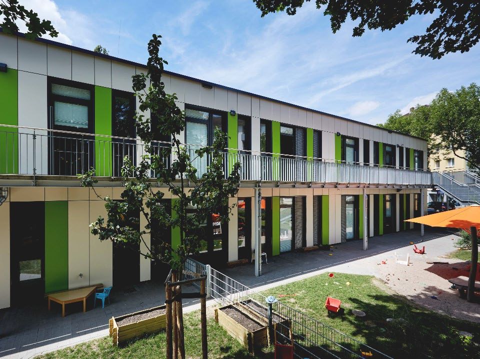 Zweistöckiges Gebäude mit grün-weißer Fassade, Balkon und Spielplatz mit Sandkasten und Sonnenschirm im Vordergrund