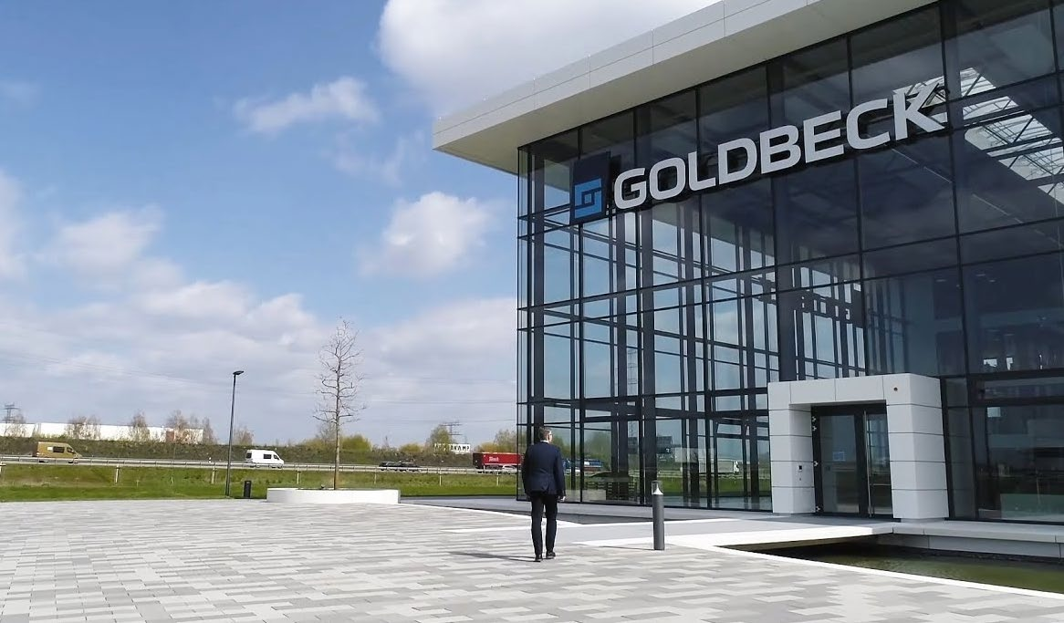 Ein Mann in dunklem Anzug geht auf ein modernes Glasgebäude mit der Aufschrift 'GOLDBECK' zu