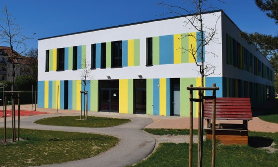 Rechteckiges zweistöckiges Gebäude mit vertikalen farbigen Streifen an der Fassade und einem kleinen Spielhaus davor