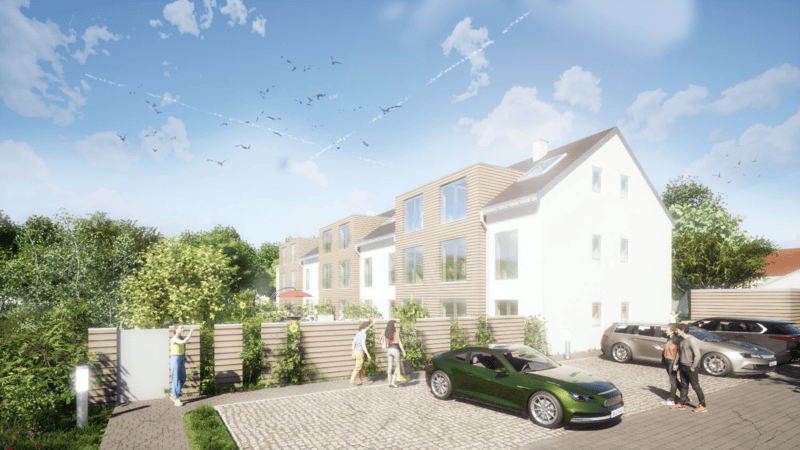 Mehrfamilienhaus mit weißer Fassade und Holzverkleidung, davor gepflasterter Parkplatz mit mehreren Autos und Menschen, umgeben von Bäumen und blauem Himmel