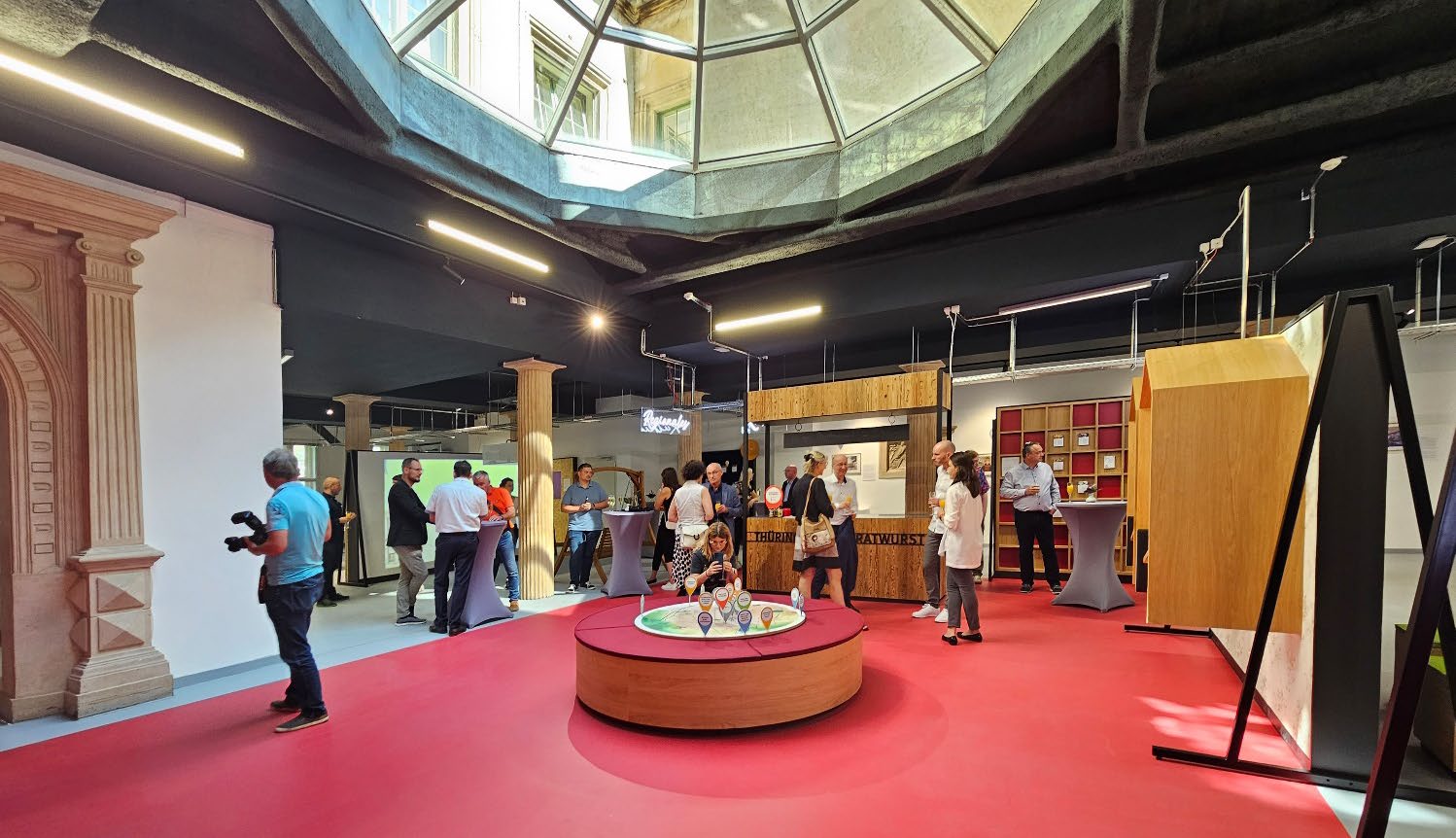 Innenraum mit rotem Boden, zentralem runden Tisch mit Dekoration, mehreren Menschen und großer Oberlichtkuppel